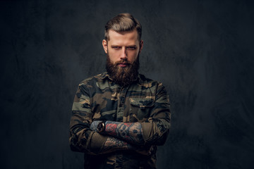 A stylish bearded guy with tattooed hands in the military shirt, posing with his arms crossed and looking sideways. Studio photo against a dark wall