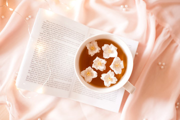 Cup of green tea with jasmine flowers staying on open book over light at background.