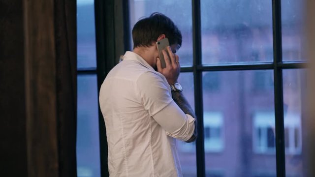 Married Caucasian man in a white creased shirt with short brown hair with watch on his hand and a tattoo talking on the beige phone. Businessman looking into a dark blue dirty wooden window. 