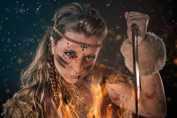 Beautiful young Amazon female warrior posing in a fighting stance with a dagger, fire flames in...