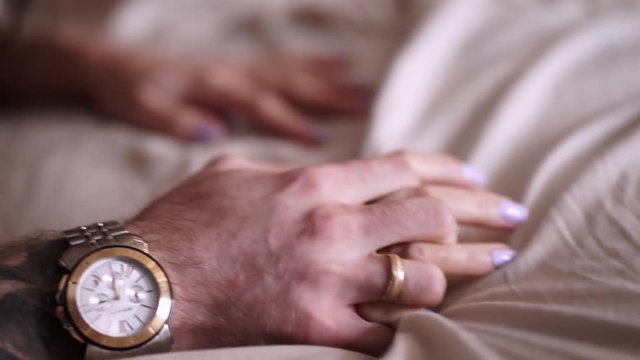 Couple in love holding hands tight, close up. Man with tattoos, watch and ring holds woman hand, selective focus. Male and female hands crossed on white cotton sheets in bed. Sex concept. 
