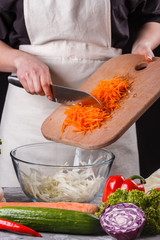 young woman in a gray apron adds carrot to a salad