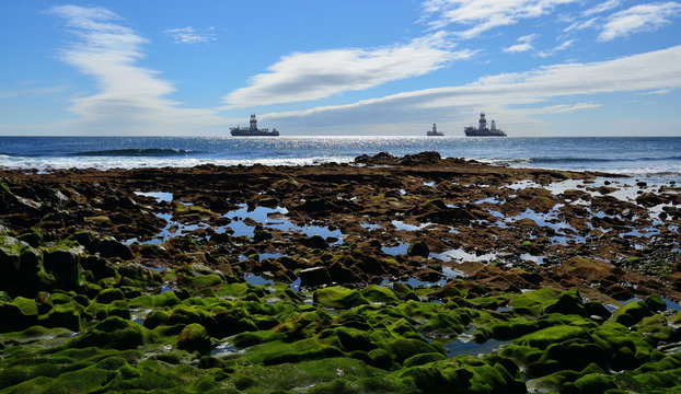 Rocky coast at low tide with many puddles, blue sky, clouds and oil platforms in the bay 