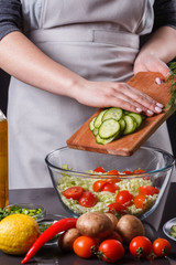 A young woman in a gray apron adds a cucumber to a salad
