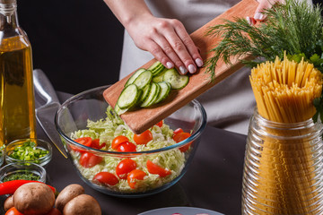 A young woman in a gray apron adds a cucumber to a salad