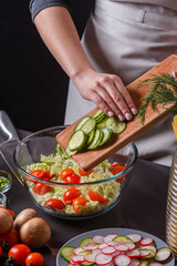 A young woman in a gray apron adds a cucumber to a salad