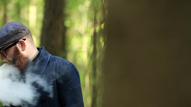 Vape bearded man. An adult white short-haired man with a very long beard in a cap and glasses smokes an electronic cigarette outside in the dark forest. Bad habit that is harmful to health.