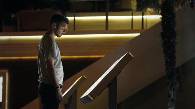 Attractive man using interactive touchscreen terminal display for searching info. Modern teen in wireless earphones. Evening time, lowlight exterior. Education and entertainment concept. Close up shot