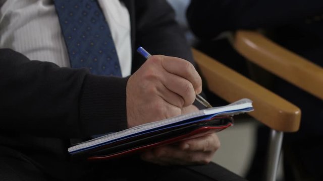 Closeup hans of middle aged marketing manager making notes with pen in notepad during speech at business conference.