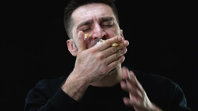 Hungry male greedily eating cake against dark background, bulimia problem