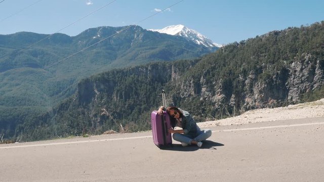 Beautiful young lady sitting exhausted on rural road with suitcase hitchhiking on sunny day outdoors
