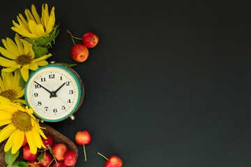 Old alarm clock near a bouquet of yellow sunflowers and flowers and red apples in a basket on a black background. Greeting card Good morning, day. The concept of summer, or September 1.