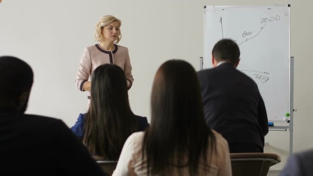 Middle aged business woman finished speech at seminar and waiting for questions from audience. Back view member of business training raising hand and asking public speaker for clarification