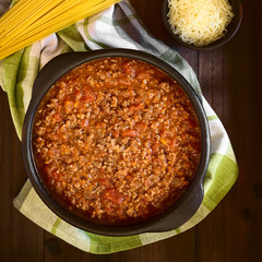 Homemade bolognese sauce made of fresh tomatoes, onion, carrot, garlic and mincemeat, uncooked spaghetti and grated cheese on the side, photographed overhead on dark wood with natural light