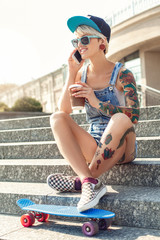 Skateboarding. Alternative girl skater in cap and sunglasses sitting on stairs foot on penny board...