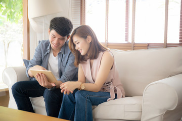 Asian young couple relax looking tablet entertainment on internet together on sofa at home, family leisure watching movie digital with comfortable, lifestyle and wellbeing concept.