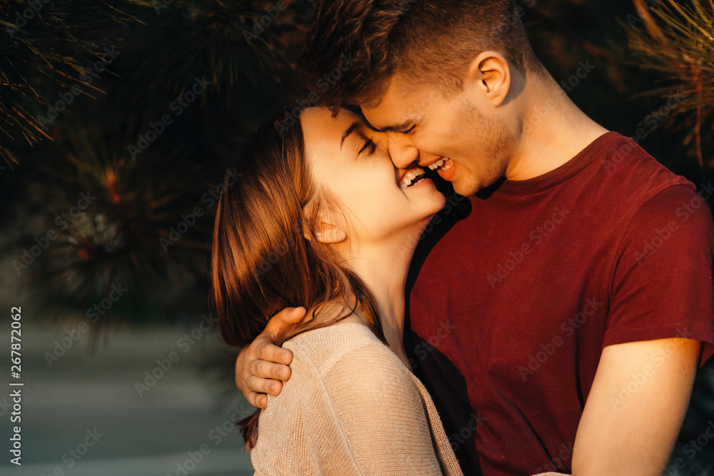 Wall mural Side view of a lovely young couple having fun smiling before kissing with closed eyes in sunset light against a tree.