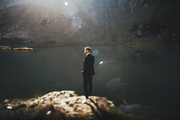 Fototapeta na wymiar Full length portrait of a handsome young caucasian man dressed in suit sitting on a rock in the mountains near a lake against sunrise looking away.