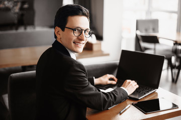 Young attractive adult freelancer with glasses looking at camera over the shoulder smiling while working at his laptop in a coffee shop.