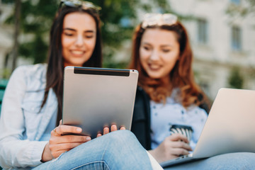 Back view of a tablet used by two lovely girls smiling sitting on a bench outside in the park.
