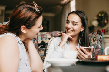 Beautiful caucasian woman with dark long hair sitting in a coffee shop smiling while listening her girlfriend with a smartphone in her hands.