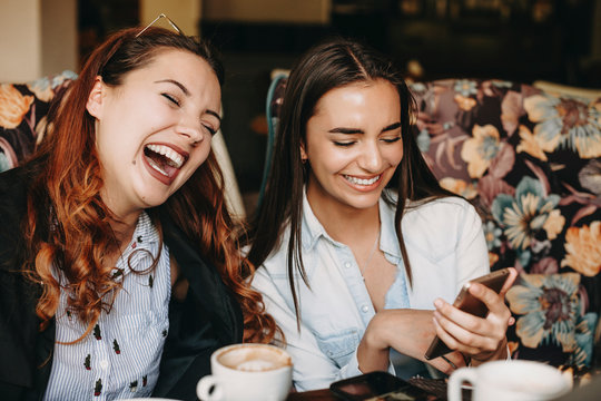Lovely young plus size woman having fun laughing with closed eyes while her girlfriend showed something on her smartphone while drinking coffee in a coffee shop.