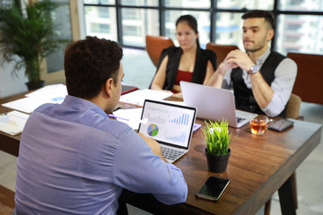 two caucasian businessmen having meeting and using computer with chart on screen and company graph summary report on table with coffee cup and calculator beside