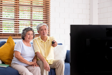 Asian couple senior sitting on sofa and use remote control to change channel and watching tv in...