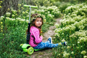 Portrait of a beautiful little girl in a wreath of daisies for a walk . Girl with a scooter for a walk in the field