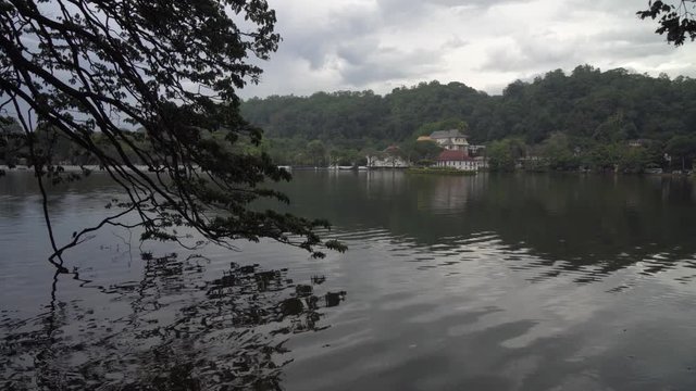 Static shot across calm lake waters & through tree branches to Sacred Buddha Tooth temple. Sri Dalada Maligawa, kandy, Sri Lanka