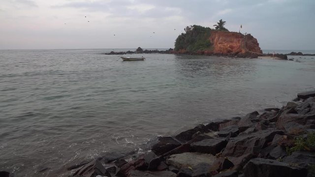 Slow panning shot across sandy, rocky section of Marissa beach, Sri Lanka. Amazing coastal large tropical rock formation emerging from seascape with boat floating below.