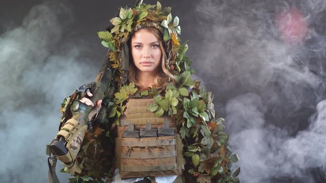Heavily armed female soldier in battle helmet and ghillie suit holding assault rifle isolated on dark smoky battlefield. Paint ball and laser tag sport games