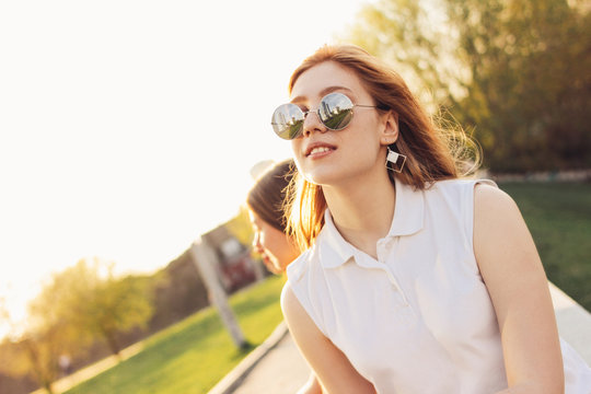Positive beautiful happy red haired girl in mirror sunglasses with friends on city street background, summer sunset time