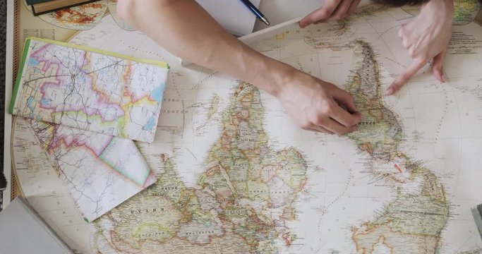 Couple man and woman are planning vacation using a world map. Pointing at the map places to visit in South Anerica using red flag pins, closeup hands.
