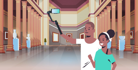 young couple using selfie stick taking photo on smartphone camera african american man woman visitors in modern art gallery museum hall interior portrait horizontal