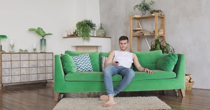 Young guy in jeans and white t-short comes in living room in his home, sits on green couch, turns on television and watching, front view.