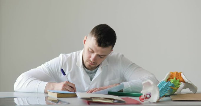 Portrait of young roentgenologist man doctor is making his paperwork writing description of the x-ray image sitting at his workplace on white background, front view.