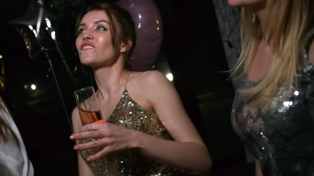 Medium tilt close-up of three relaxed young dressed-up female friends partying together, enjoying champagne and making fake moustache with each other&rsquo;s hair