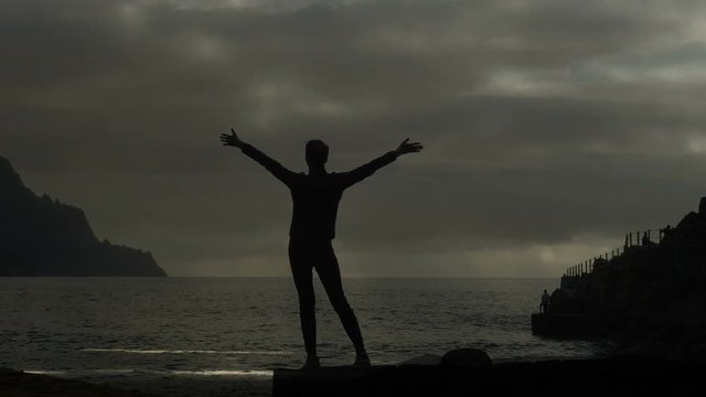 Silhouette of a woman at sunset raising arms in the air observing waves and dramatic thunderstorm clouds. Time-lapse of fastmoving dark storm clouds