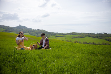 A young couple in bright clothes sitting on a bright green meadow and drinking wine
