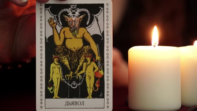 A witch is fortune teller in mantle shows tarot card. White candles on table. Occult, esoteric, divination and wicca concept. Translation: The Devil.