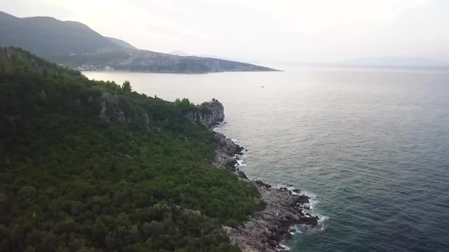Flying along the coastline with waves crashing into the rock from the Adriatic Sea. Sunrise over Himara village along the beautiful Albanian Riviera during the summer