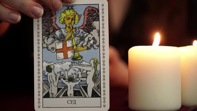 A witch is fortune teller in mantle shows tarot card. White candles on table. Occult, esoteric, divination and wicca concept. Translation: The Judgment.