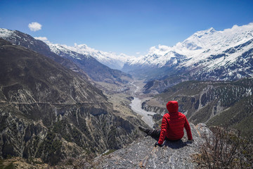 Explorer concept: Man wearing red jacket seating on the edge of a clift in Thorong-La Base Camp located in Annapurna Circuit