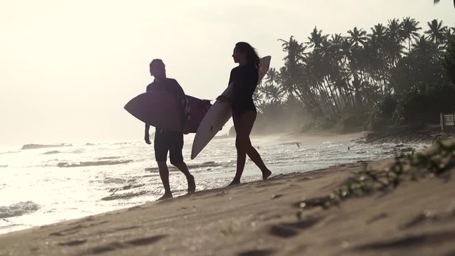 Two young people are walking with a surfboard on the beach at the sunset time. A romantic couple of surfers.