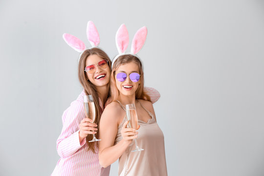 Beautiful young women in pajamas and with party decor drinking champagne on light background