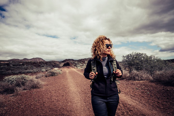 Beautiful trekker caucasian blonde curly young active woman walk in the desert path enjoying the freedom and the active lifestyle traveling with backpack alone in the outdoors natural park