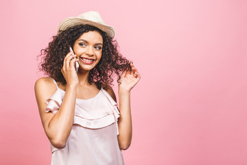Portrait of a cute happy afro american girl in dress talking on mobile phone and laughing isolated over pink background.