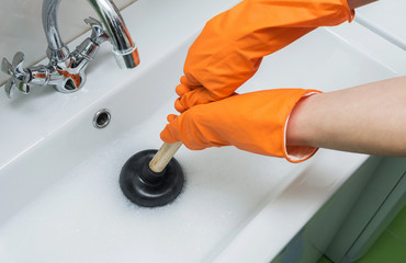 Person in protective orange gloves unblocking a clogged sink with plunger or rubber pump. Close-up,...