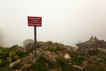 A red warning sign on the edge of a cliff with the words "Attention! Do not come to the rock! Dangerously!"